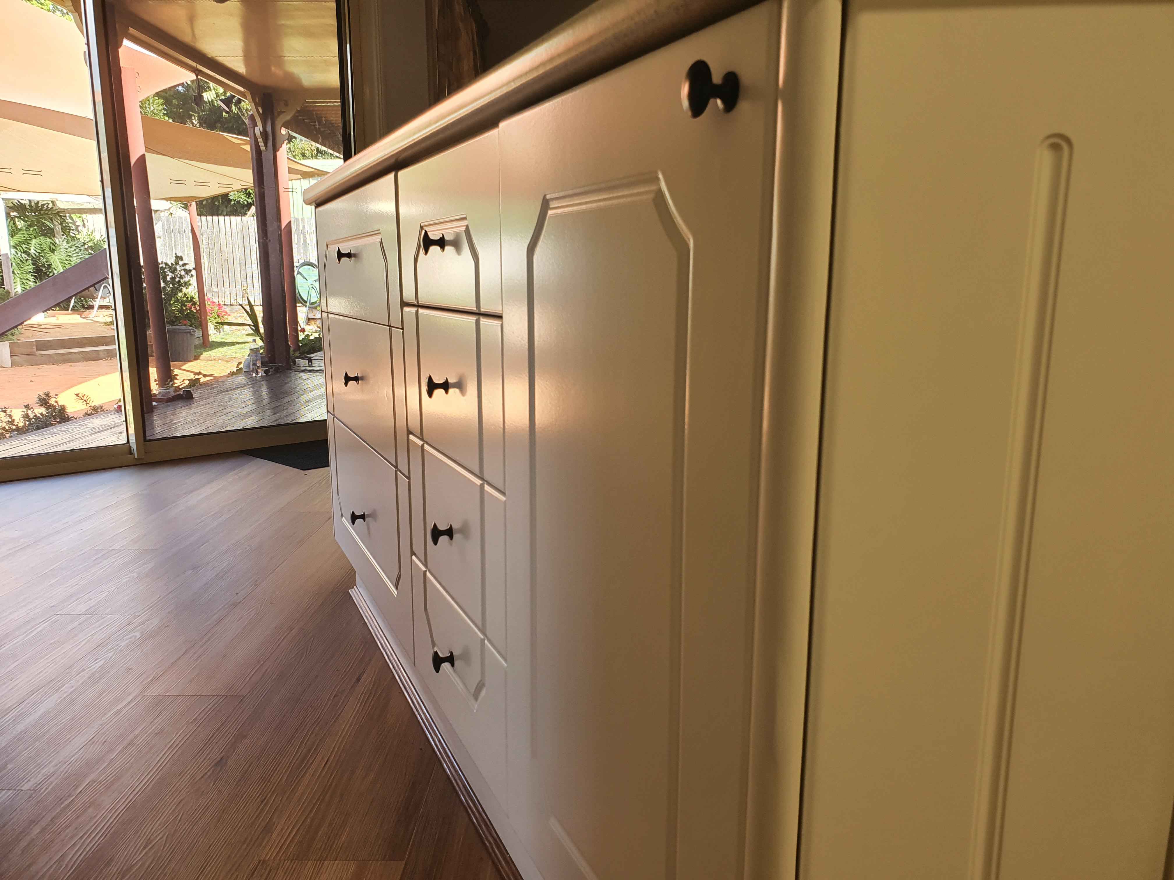 White dresser with many drawers and black knob handles viewed from the indoors, with a glass door and patio outside to the left.