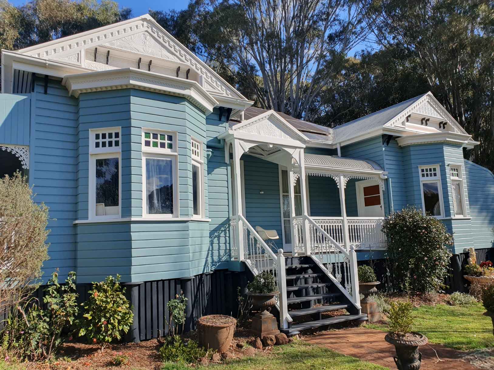 residential_painting Blue wooden Victorian-style house with white trim and ornate porch, set among trees in a sunny yard.